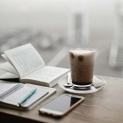 A sweet coffee, notebook, phone, and book sit on a wooden desk with a blurred view out the window