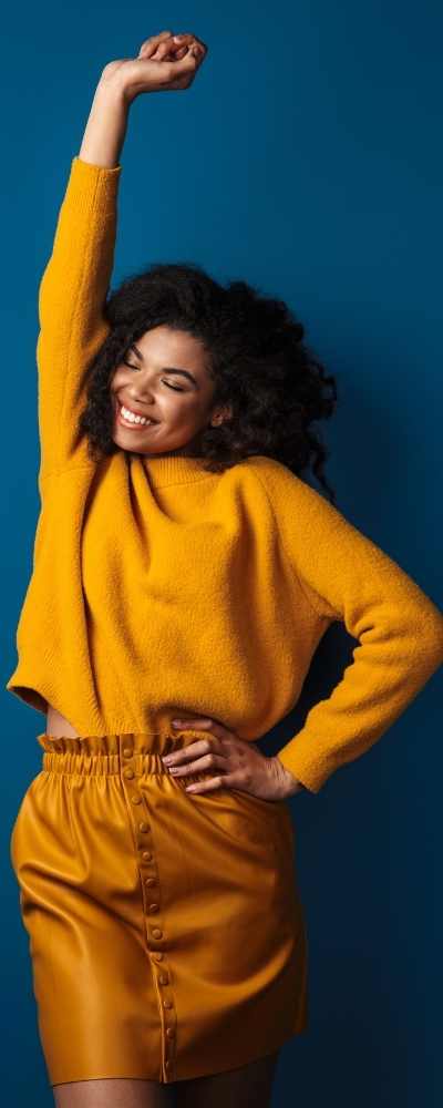 A Black woman in orange stands smiling with right arm raised up and left on her hip against a blue wall A Black woman in orange stands smiling with right arm raised up and left on her hip against a blue wall