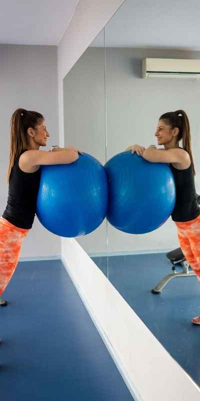A woman leans on a blue exercise ball while smiling to herself in the mirror after a workout feeling proud of herself for committing to exercise despite her executive function challenges