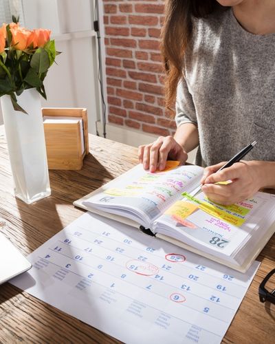 A woman writes in an agenda book with a calendar on the desk above, using post it notes and color coding to help with her executive functioning challenges
