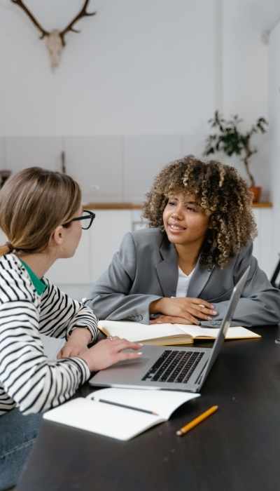 Two women sit at a table having a conversation with their laptop and notebooks in front of them Two women sit at a table having a conversation with their laptop and notebooks in front of them