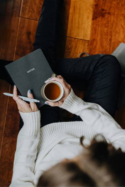 A woman holds a mug of tea with a notebook and pen preparing to reflect on the prompts about letting go