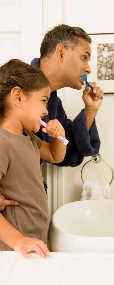 An adult and child standing together at a sink and brushing their teeth together