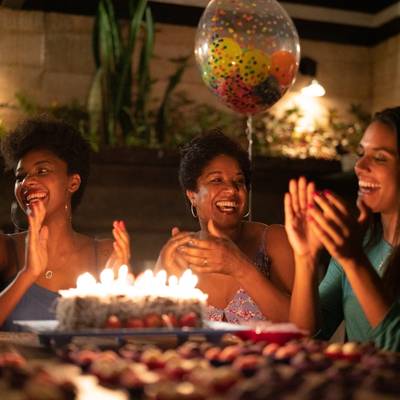 A group of adult women around a birthday cake with lit candles clap and celebrate together as part of an annual get-together