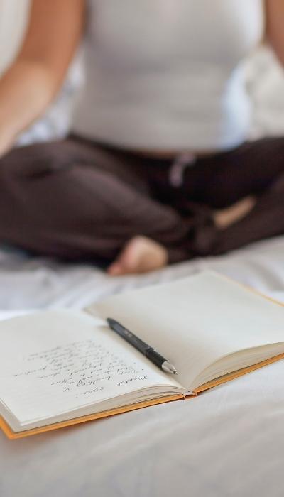A woman sits cross-legged on the bed doing a breathing exercise with her journal open on the bed in front of her