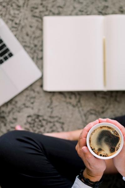 A notebook with pen beside a laptop as someone sits holding a coffee in front of them on the floor